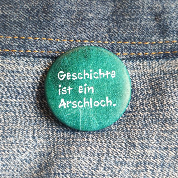 Ansteckbutton Geschichte ist ein Arschloch auf Jeans